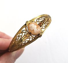 "Antique art deco cameo bracelet. The hinged bangle bracelet is a gold wash over ornate stamped metal and filigree. A small pink shell cameo of a woman's profile is the prong set focal. The unusual clasp features a very long tongue that slides along the interior of the bracelet. Size: Interior diameter of the bracelet is 7\". The width is 3/4\" at its widest and 1/4\" at its narrowest. The cameo is 1/2\" x 3/8\". Condition: Excellent antique condition. The clasp opens and closes securely. There Vintage Rose Gold Jewelry With Intricate Design, Gold Cabochon Bangle Jewelry, Ornate Cameo Jewelry For Formal Occasions, Elegant Adjustable Cameo Bracelets, Antique Gold Cabochon Bracelets, Antique Gold Bracelets With Cabochon, Victorian Gold Bracelets With Cabochon, Adjustable Gold Cameo Jewelry, Victorian Gold Cuff Bracelet Gift