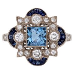 Ring in 18 karat white gold. This Art Deco-style ring is set with a square aquamarine in millegrain closed setting in the center of an openwork design set with modern brilliant-cut diamonds in millegrain setting. At each cardinal point of the setting, a half-moon motif is adorned with 5 calibrated sapphires. On either side of the head, at the start of the ring, 2x3 modern brilliant-cut diamonds are set. The basket is openworked. Total weight of the sapphires : 1,10 carat approximately. Total wei Blue Square-cut Platinum Ring, Blue Platinum Rings With Square Cut, Blue Square Cut Platinum Ring, Blue Platinum Square Cut Ring, White Gold Sapphire Ring With Square Cut, Blue Multi-stone Sapphire Ring In Art Deco Style, Luxury White Gold Topaz Ring With Multi-stone, Luxury White Gold Multi-stone Topaz Ring, Luxury Multi-stone White Gold Topaz Ring