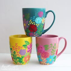 three colorful coffee cups sitting next to each other on a white counter top and one has flowers painted on it