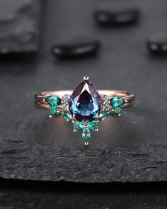 an engagement ring with blue and green stones on top of a black stone slab in front of