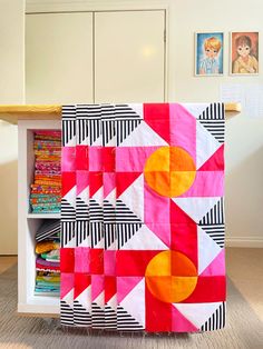 a colorful quilt sitting on top of a white book shelf next to a wooden table