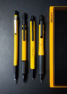 four different pens are lined up next to each other on a black table top with a yellow case