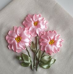 three pink flowers with green leaves are on a white linen material piece that has been embroidered onto