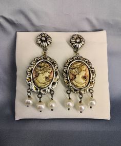 Silver dangle earrings with handmade ceramic cameo with three dangling pearls, Victorian style. These earrings are very original jewels, they reflect the classic shape of the cameos that we can admire in ancient portraits. The cameo is handmade in ivory-colored synthetic ceramic on a light pink background typical of shell cameos. These earrings are made entirely by hand in all their parts and silvered with a manual procedure with antiqued and vitrified metal leaf which makes them shiny and protects them. This jewel is shipped with registered shipping and is packaged in an elegant blue velvet and white satin gift box, very suitable as a gift, and the package also includes an elegant description of the jewel printed on straw-coloured paper. When placing the order, together with your shipping 18th Century Jewelry Earrings, Elegant Cameo Earrings, Victorian Cameo Earrings Collectible, Collectible Cameo Pendant Brooches, Ornate Cameo Necklace Collectible, Cameo Earrings, Ivory Color, Silver Earrings Dangle, Pink Background