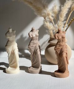 three ceramic sculptures sitting next to each other on a white tablecloth with pamodia in vases behind them