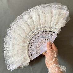 a hand holding a white lace fan on top of a gray sheet covered tablecloth