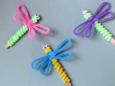 three dragonflys made out of colored yarn on a gray surface, one is yellow and the other is purple