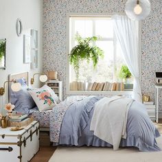 a bed room with a neatly made bed next to a window filled with books and plants