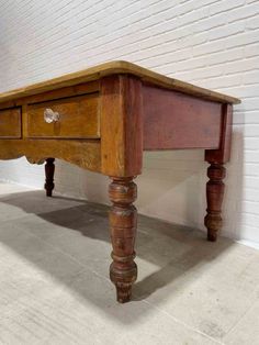 an old wooden table with two drawers on one side and three legs, in front of a white brick wall