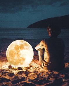 a person sitting in the sand next to a full moon