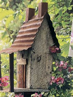 a bird house sitting on top of a wooden post next to pink flowers and trees