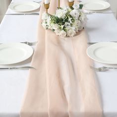 the table is set with white plates, silverware and pink cloth draped over it