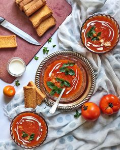 two bowls filled with tomato soup next to bread and tomatoes on a tablecloth,