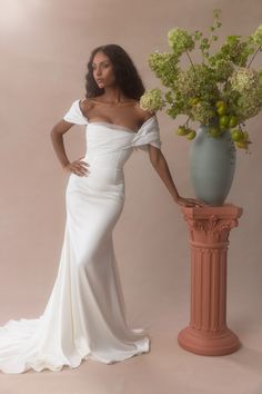 a woman standing next to a vase with flowers in it and wearing a white dress