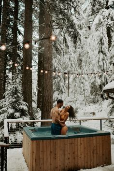 a man and woman are in the hot tub surrounded by snowy trees with lights strung above them