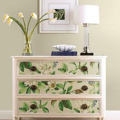 a white dresser with flowers on it next to a lamp