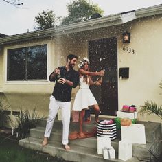 a man and woman standing on the steps in front of a house holding wine glasses