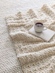 a cup of coffee sits on top of a white crocheted blanket with a book