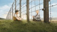 two children are sitting on the grass near a fence with barbed wire and looking at each other