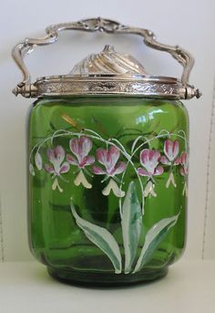 a green glass jar with pink flowers painted on the front and silver trimmings