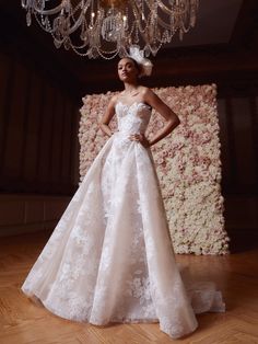 a woman standing in front of a chandelier wearing a wedding dress with flowers on it