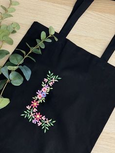 a black tote bag with pink and purple flowers on it next to a green plant