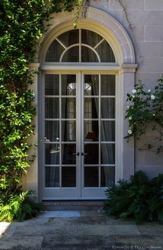 an arched window with white curtains on the side of a building next to green plants