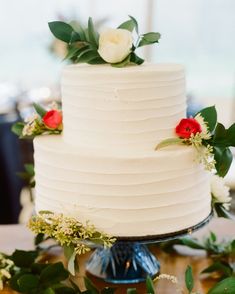 a white wedding cake with red and white flowers on the top is surrounded by greenery