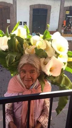 an old woman with flowers on her head is standing by a fence and looking at the camera