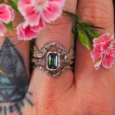 a close up of a person's hand with a ring on it and flowers in the background