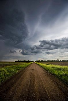 a dirt road in the middle of a field under a dark sky with storm clouds