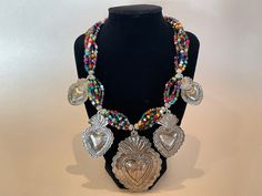 "Striking Mexican Costume Jewelry with Five Large Sacred Hearts (in three sizes). Seven strands of multicolor glass beads hang from starts. Strands are grouped into six sections by larger metal beads. At each of these sections is a Sacred Heart. Large TIN Sacred Heart (3 1/2\" long) hangs from bottom of glass beads. Two pairs of smaller identical Sacred Hearts hang close by. Necklace is of a medium length, 12\".  Costume Jewelry is completely HANDMADE, as a result, there may be subtle imperfections. These small variations enhance the value and beauty of each piece. We are proud of the level of quality of this item." Traditional Multicolor Heart Beads Necklace, Multicolor Heart Beads Necklace For Festivals, Artisan Multicolor Heart-shaped Jewelry, Traditional Multicolor Jewelry With Heart Beads, Multicolor Heart Beads Jewelry For Festival, Multicolor Spiritual Jewelry With Heart Beads, Spiritual Multicolor Beaded Necklaces With Heart Beads, Festival Multicolor Heart Beads Jewelry, Multicolor Heart Jewelry For Festivals