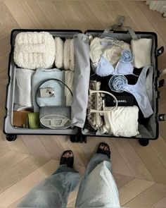 an open suitcase filled with clothes and other items on top of a wooden floor next to someone's feet