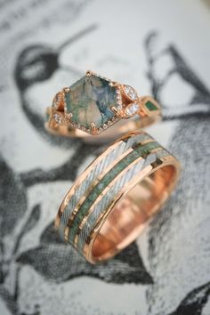 A matching wedding ring set from Staghead Designs sits on an antique bird illustration. First is our "Lucy In The Sky" design engagement ring in 14K rose gold with a hexagon cut moss agate center stone, diamond halo & accents, & moss inlays. Below is "Rio" in 14K rose gold with a centered crushed moldavite inlay & a damascus steel inlay on either side. These rings are fully customizable & can be made with a wide variety of unique materials & stones. Make Wedding Rings, Rings Asthetic Picture, Gold Stone Rings For Women, Moldavite Engagement Ring, Womens Engagement Rings Unique, Mens Ring With Stone, Unique Engagement Rings Color, Miss Agate Wedding Ring, Hexagon Gemstone Ring