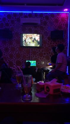 two people sitting at a table watching tv in a room with purple lighting and wallpaper