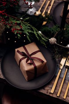 a christmas table setting with a wrapped present on a black plate and gold utensils