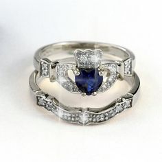 two wedding rings with blue and white stones in them on a white surface, the ring has a heart shaped sapphire surrounded by smaller diamonds