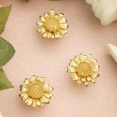 three gold flower studs sitting on top of a pink surface