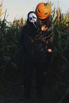 two people wearing masks standing in a corn field with pumpkins on their heads and one holding the other