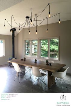 a dining room table with white chairs and lights hanging from the ceiling above it in front of two windows