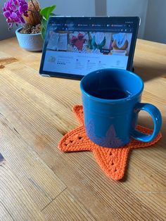 a laptop computer sitting on top of a wooden table next to a blue cup and orange crocheted coaster