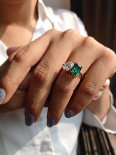 a woman's hand holding an emerald and diamond ring on her left hand, while wearing a white shirt