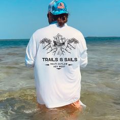 a man standing in the ocean wearing a white shirt and blue hat that says trails & sails