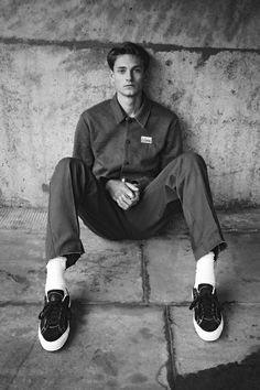 a young man sitting on the ground with his feet crossed and wearing sneakers, in front of a concrete wall