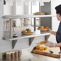 a woman in an industrial kitchen holding plates with food on the trays behind her