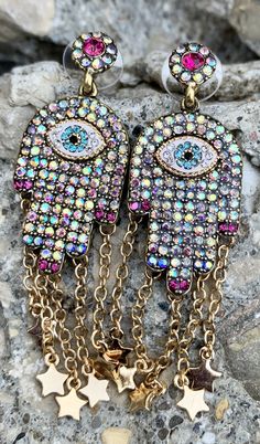 Betsey Johnson Evil Eye Multi-Color Stone Hamsa Hand Earrings Gold Tone Plating Gold Tone Earrings Covered in Iridescent Faceted Stones Featuring a Center Blue Evil Eye with Chain Drops and Stars. Multi-Color Cubic Zirconia Post Back Closure with Discs Approximate Measurements: Drop- 3" The Hamsa Hand is an ancient Middle Eastern amulet symbolizing the Hand of God. In all faiths it is a protective sign. It brings its owner happiness, luck, health, and good fortune. Eye-catching Multicolor Jewelry For Party, Multicolor Jeweled Drop Earrings, Multicolor Jeweled Metal Earrings, Eye-catching Multicolor Party Jewelry, Multicolor Metal Dangle Chandelier Earrings, Rainbow Metal Earrings With Ear Wire, Rainbow Dangle Metal Earrings, Multicolor Dangle Plug Earrings With Matching Set, Multicolor Jeweled Chandelier Earrings As Gift