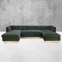 a green velvet sectional sofa with gold legs and foot rests on a concrete floor in front of a gray wall