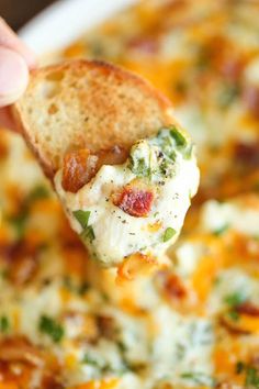 a hand holding a tortilla chip over a casserole dish filled with cheese and bacon