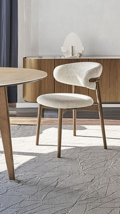 A modern Benenden Boucle Dining Chair situated in a room with a minimalist aesthetic. The chair features a textured beige linen upholstery with a gently curved backrest and a comfortable, rounded seat. It stands on four sleek, black wooden legs, positioned on a light wood-grain floor, complementing a dark dining table partially visible in the background. The room is bright, with sheer white curtains diffusing light from a window, adding to the chair’s inviting and sophisticated presence. Soft Beige, Contemporary Chic, Elegant Interiors, Understated Elegance, Modern Dining