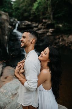 a man and woman standing next to a waterfall in the forest smiling at each other